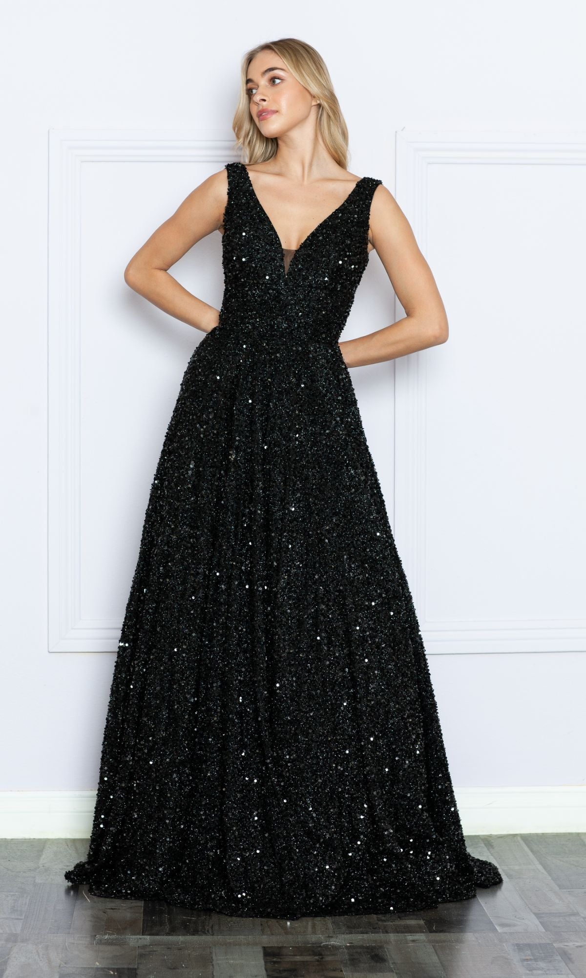 Woman wearing the Long Black Sequin A-Line Prom Dress 9168, a sleeveless, floor-length gown with a deep V-neckline, standing against a plain white wall.